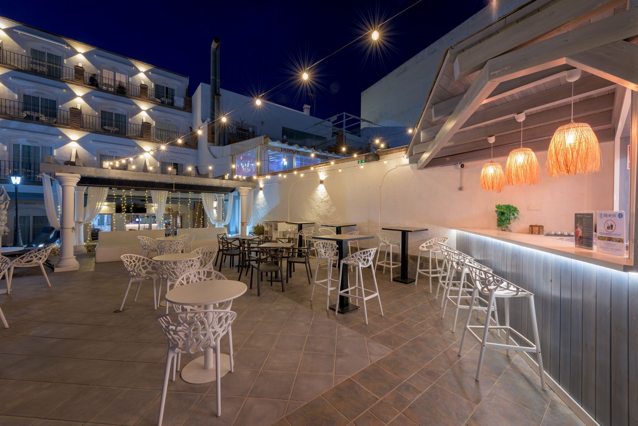 An outdoor dining area with white chairs and tables, string lights, and a bar area with a counter.