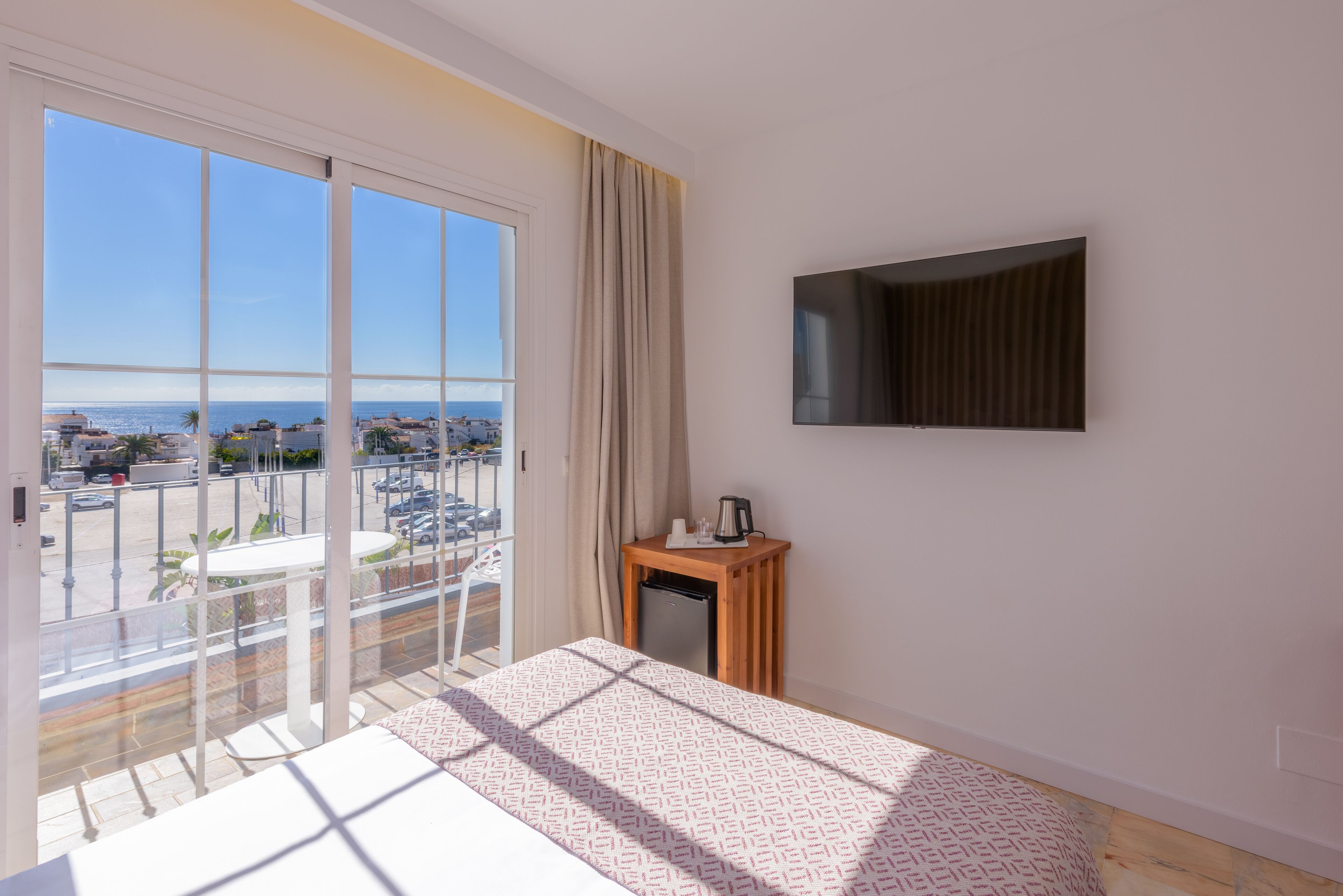 Una habitación de hotel con balcón, una cama, una mesita con hervidor y un televisor en la pared.