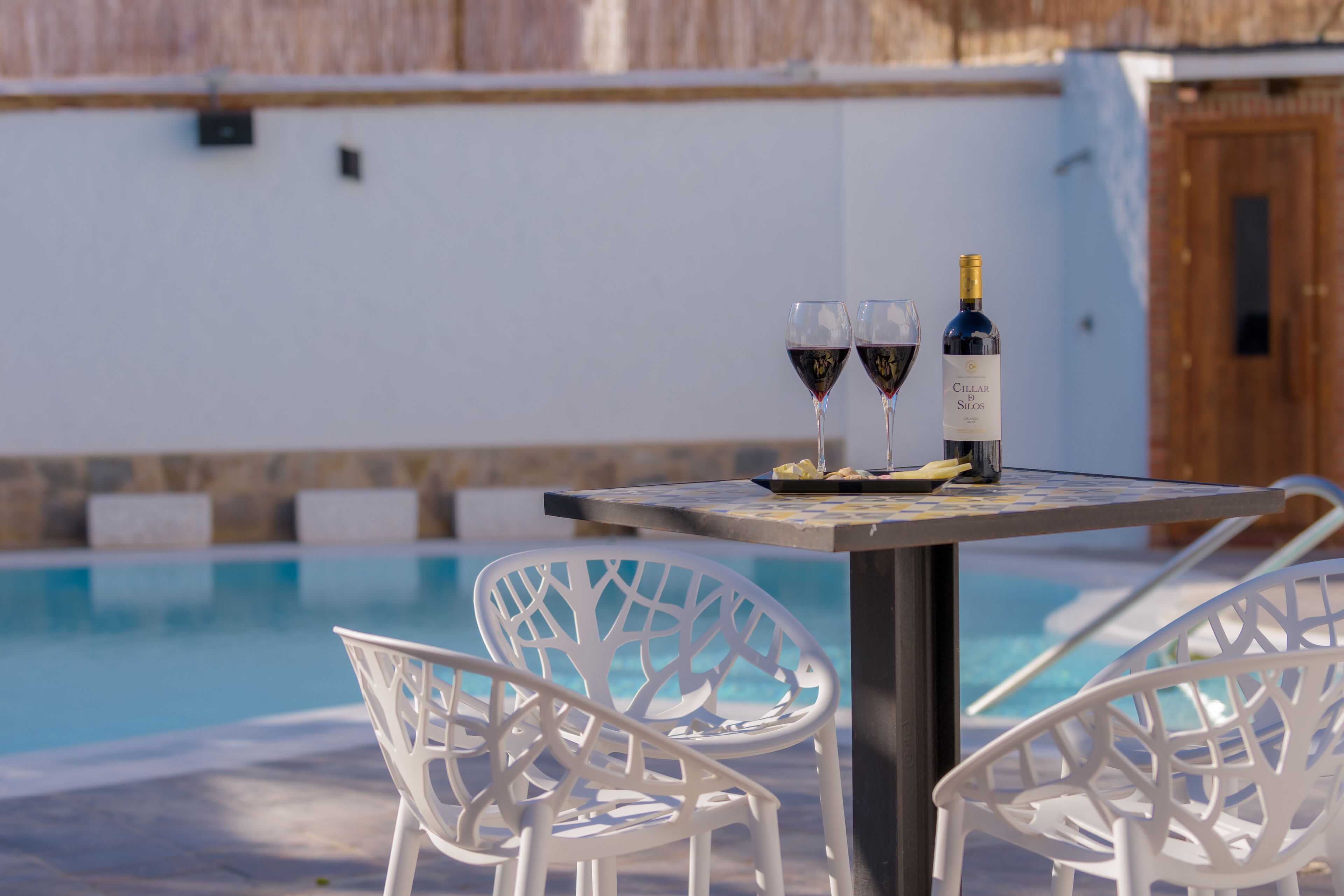 Une table placée au bord de la piscine, avec deux verres à vin et une bouteille de vin, entourée de chaises blanches.