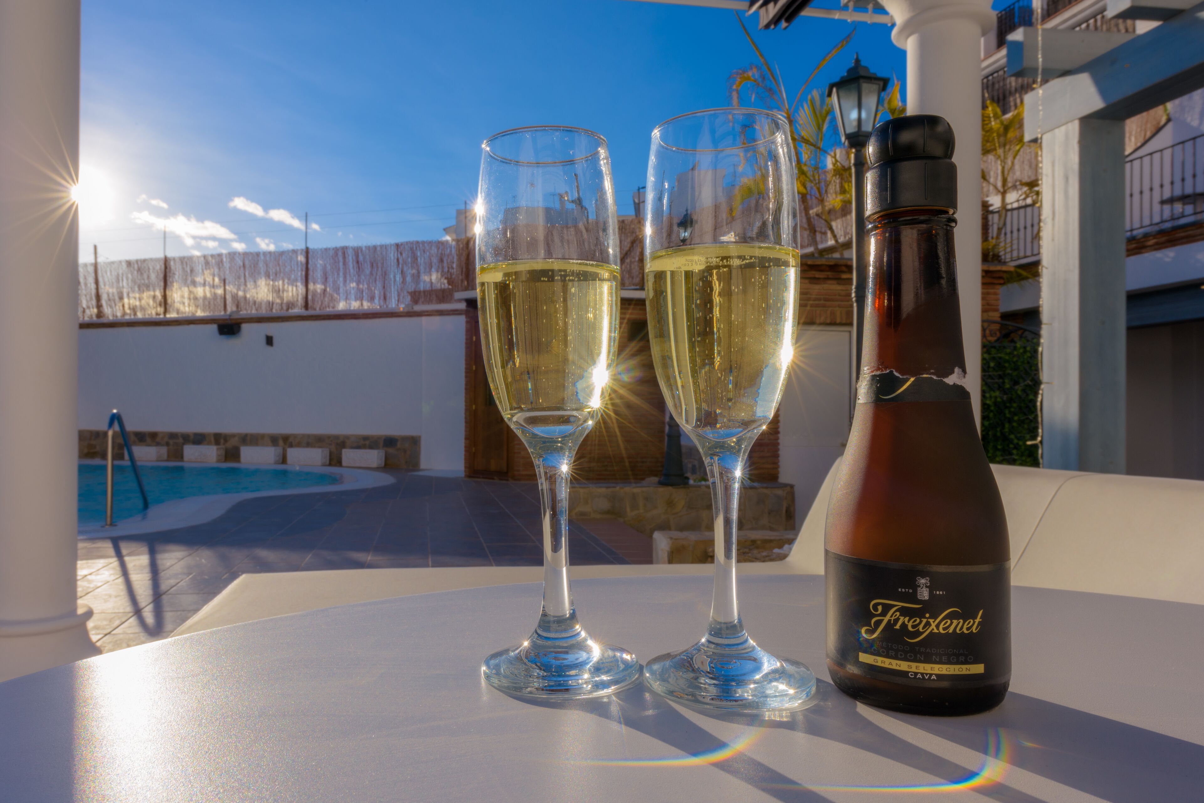 Two glasses of white wine and a bottle of Freixenet Cava on a table with a pool in the background.