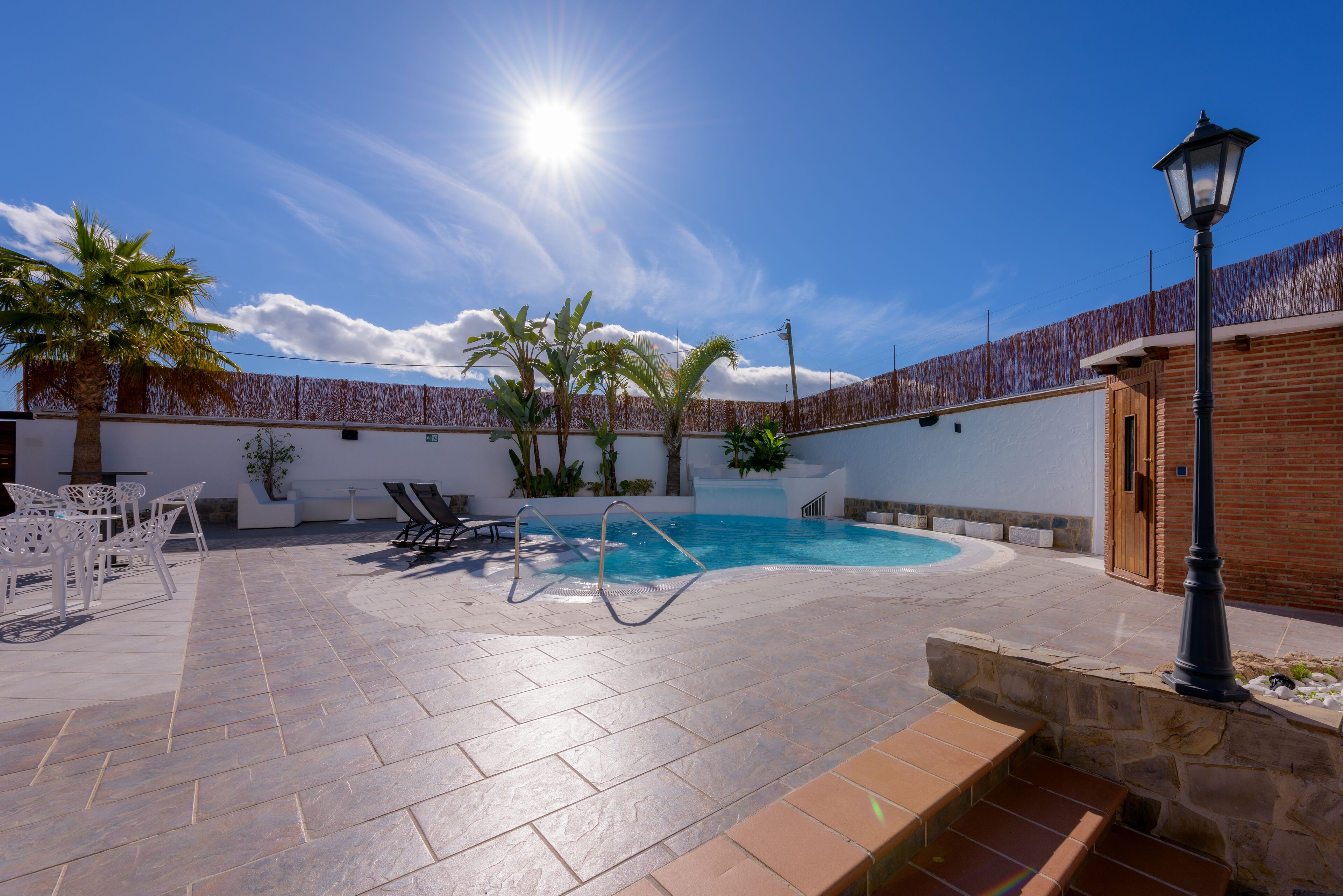 A swimming pool with a sunlit sky, a covered patio area with chairs, and a lamp post.