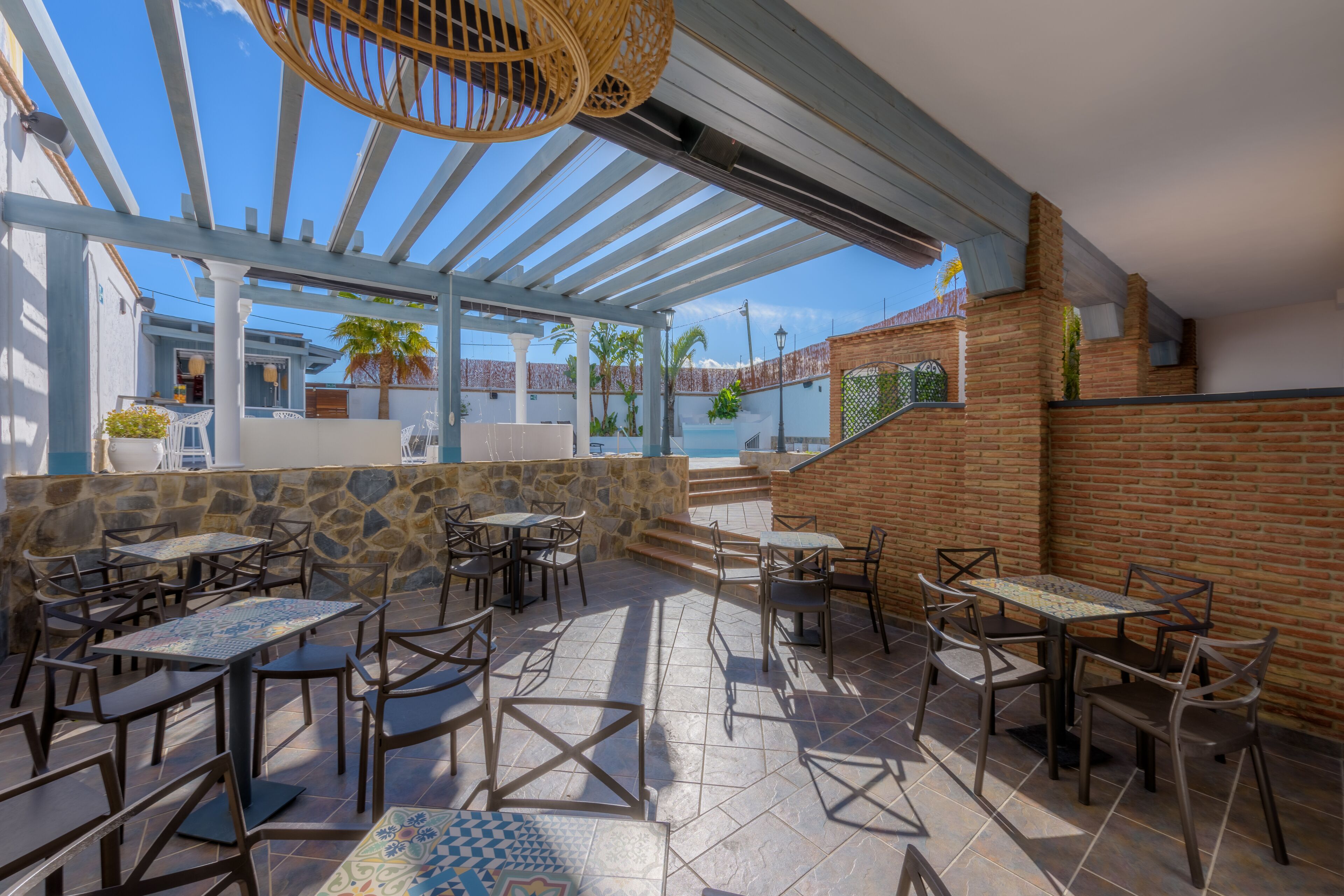 Una terrazza esterna con tavoli e sedie, un muro in pietra e vista su una piscina e degli edifici sullo sfondo.