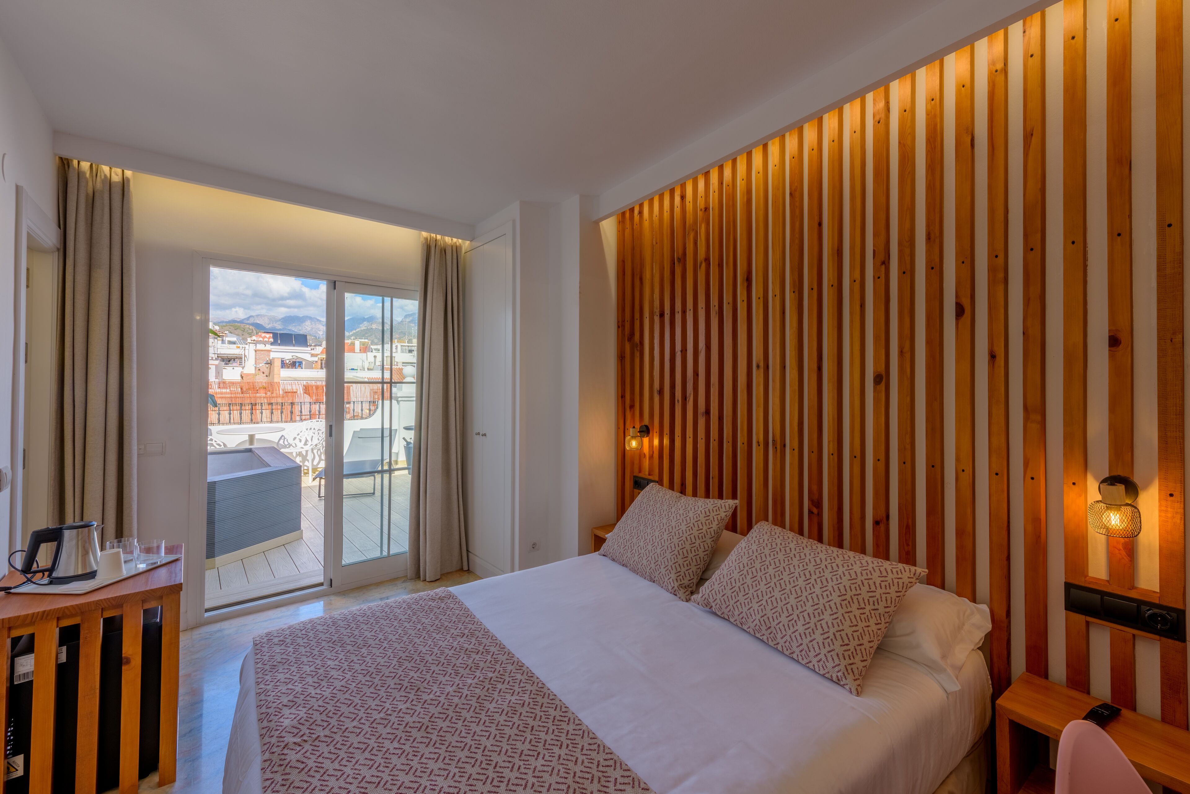 Et hotelværelse med en stor seng, en trævæg som detalje, et skrivebord med elkedel og udsigt til byen gennem en skydedør.