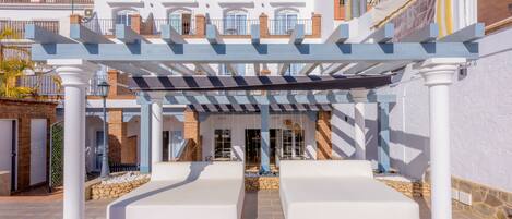 Un área de estar al aire libre con techo, columnas blancas y una pérgola, que conduce a un edificio con varios balcones y ventanas.