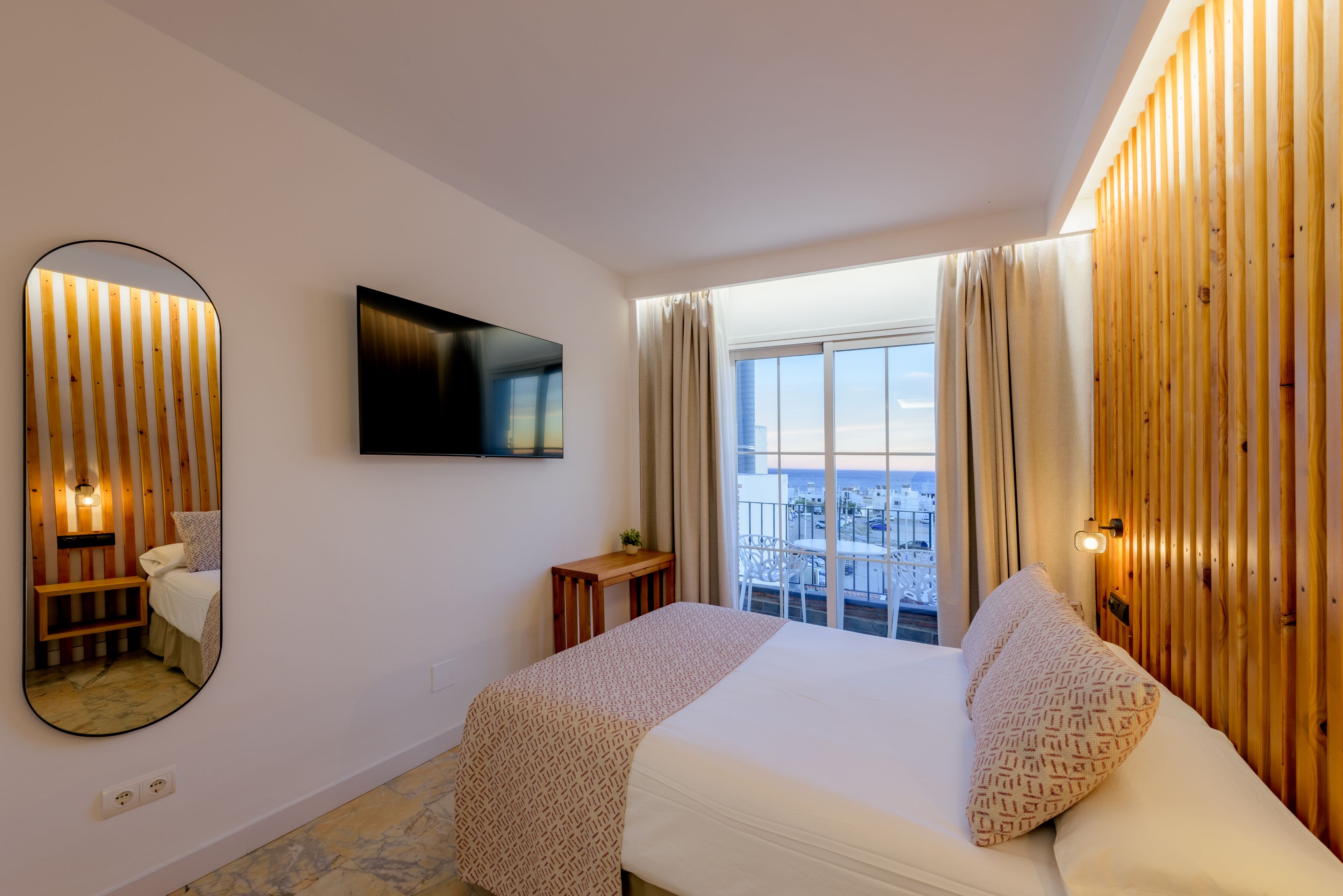A hotel room with a bed, a TV, a large window with curtains, a wooden wall panel, and a small table with a plant.