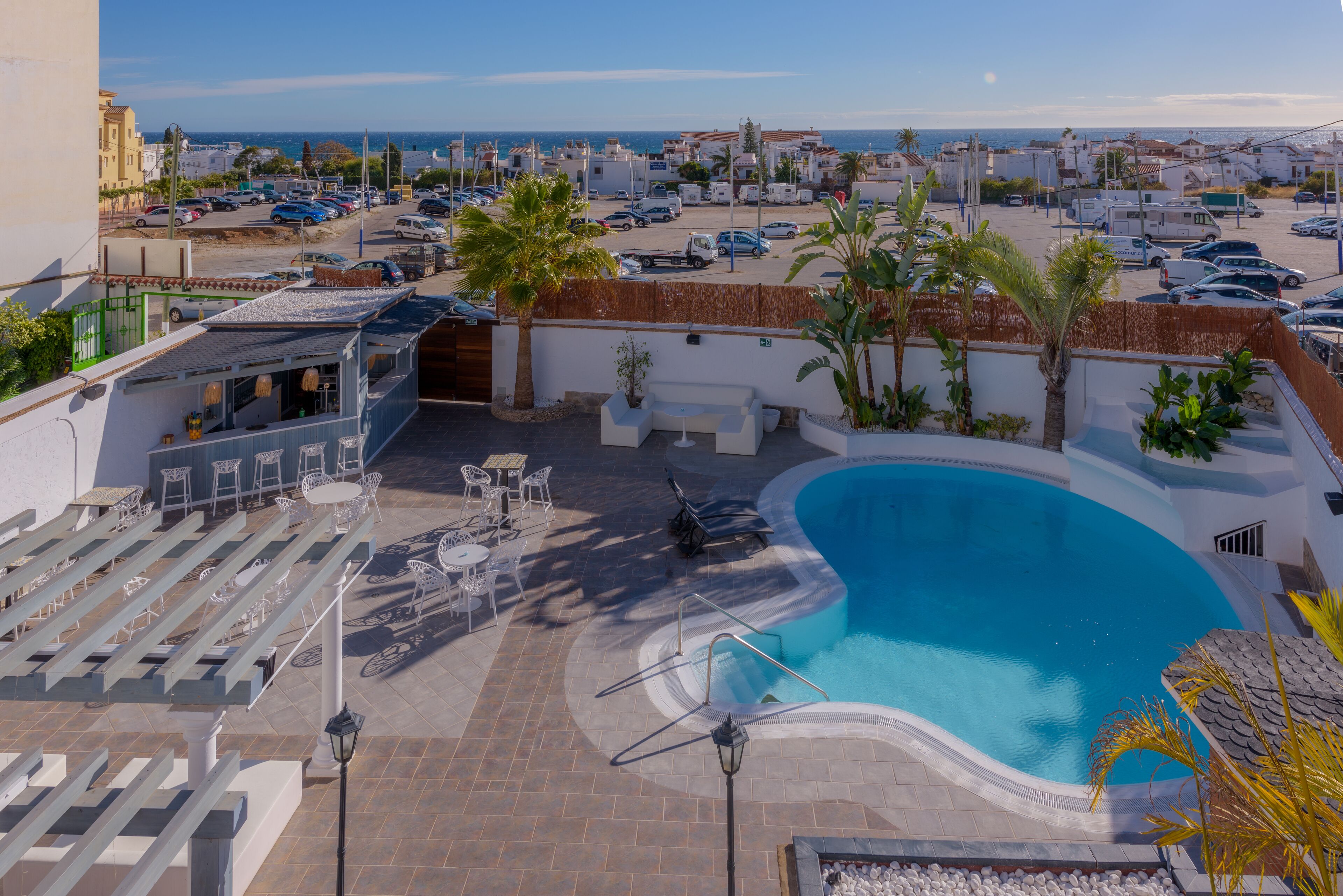 Un área de piscina en la azotea con vista al mar, mobiliario exterior y un estacionamiento.