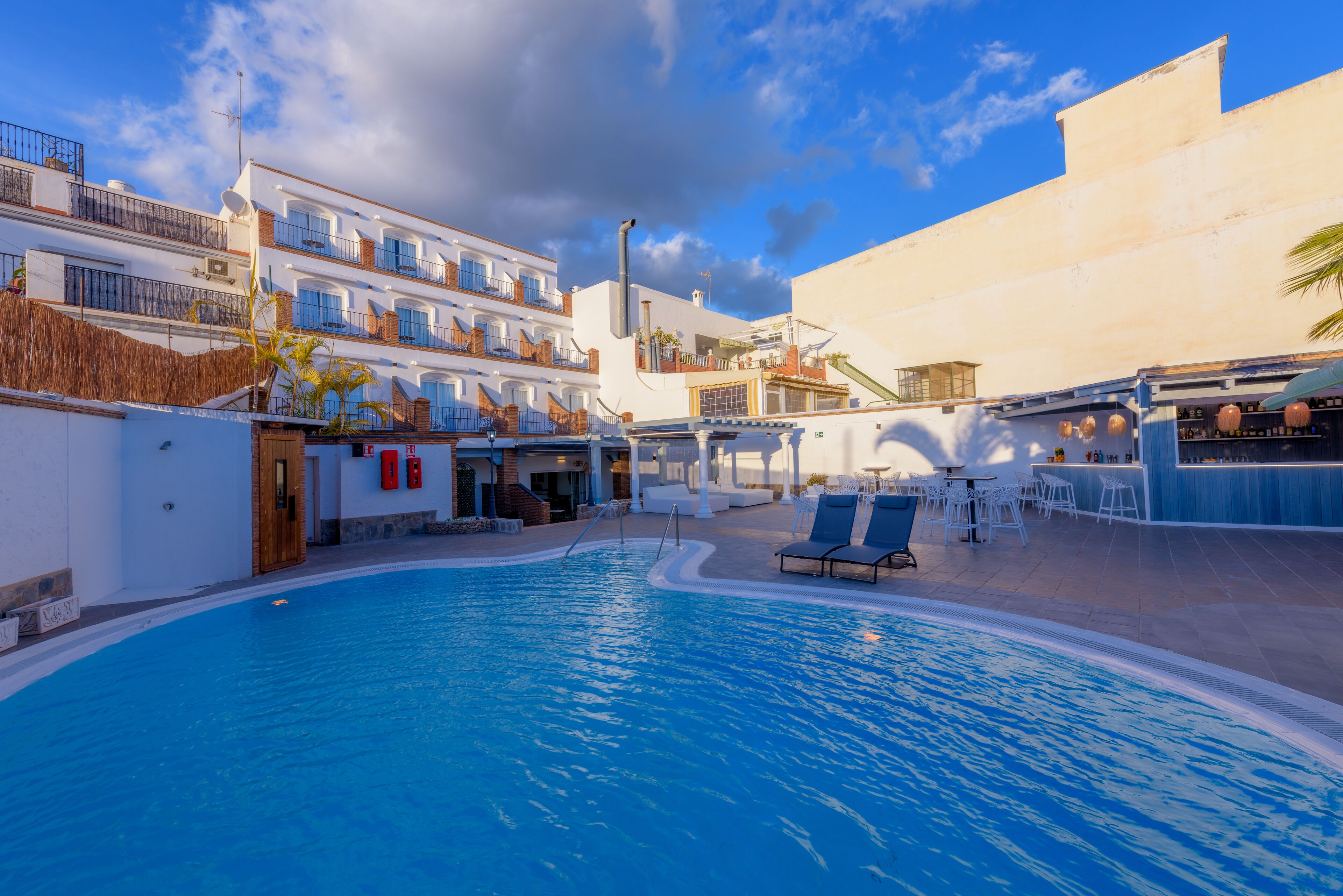 A swimming pool with lounge chairs and a bar area, surrounded by buildings with balconies and a clear sky.