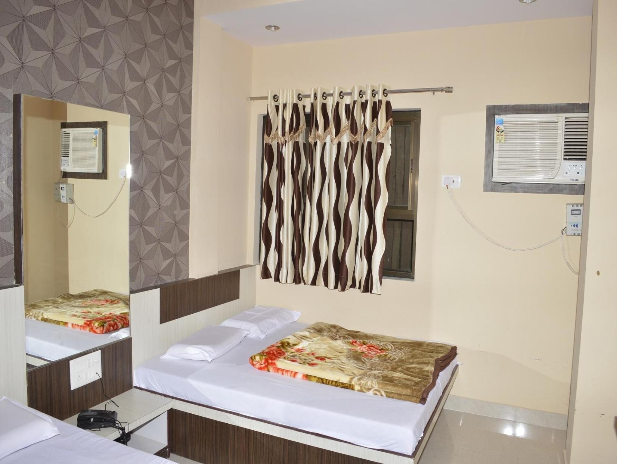 A hotel room with a bed, a mirror, a window with curtains, an air conditioning unit, and a wall-mounted control panel.