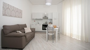 A modern living room with a brown sofa, a dining table with chairs, and a kitchen area in the background. - Aria Di Mare B&b (Marsala)