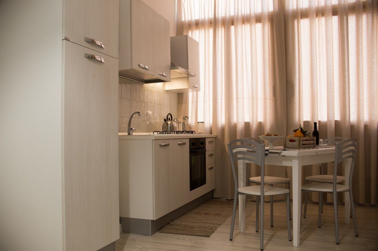 A modern kitchen with a built-in oven, a gas stove, and a range hood. There is a dining table with chairs and a window with sheer curtains.