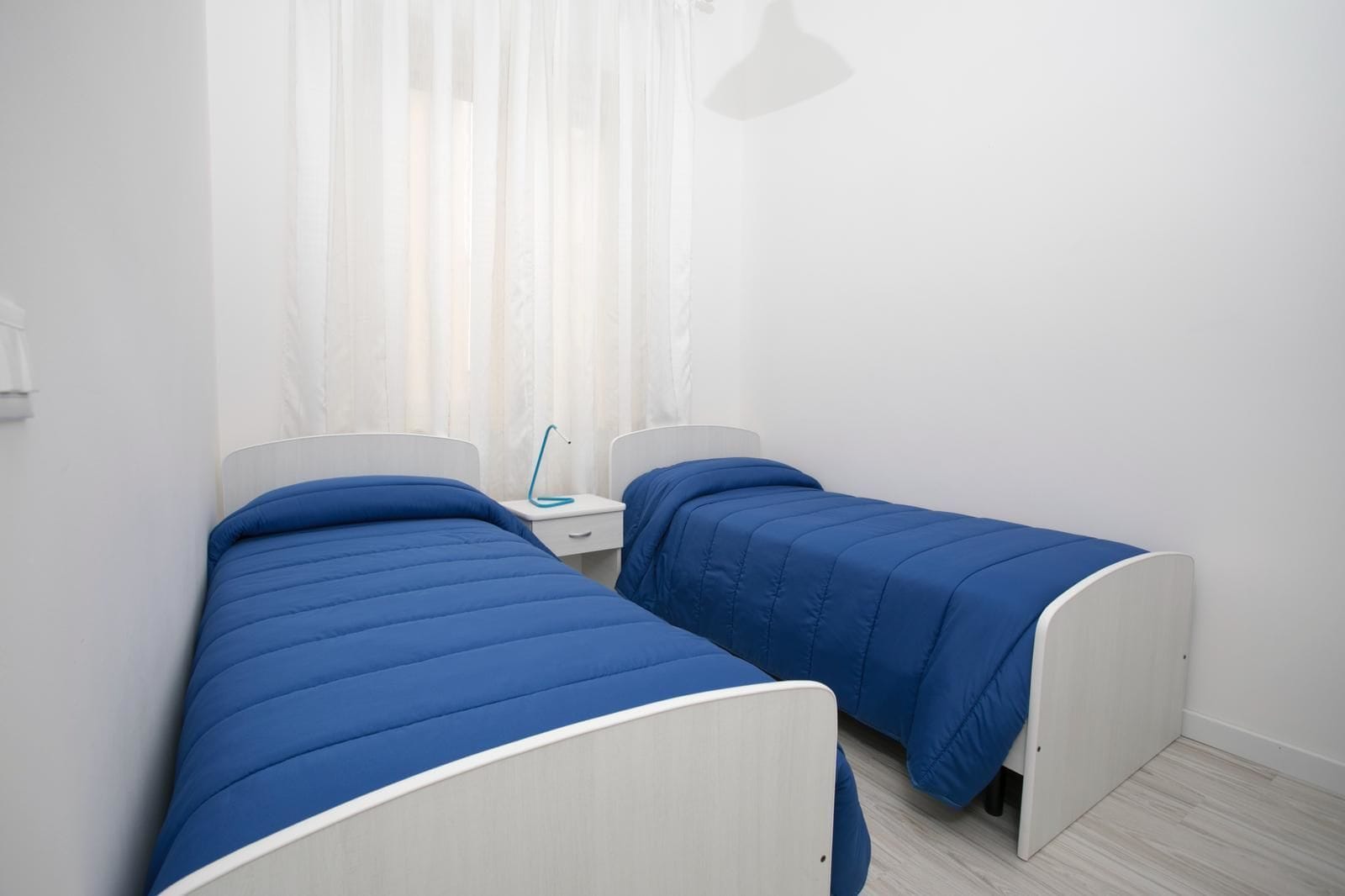 A room with two single beds, each with a blue bedspread and white headboards, a bedside table with a lamp, and a window with sheer curtains.