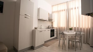 A modern kitchen with white cabinetry, a dining area with a table and chairs, and a window with curtains. - Aria Di Mare B&b (Marsala)