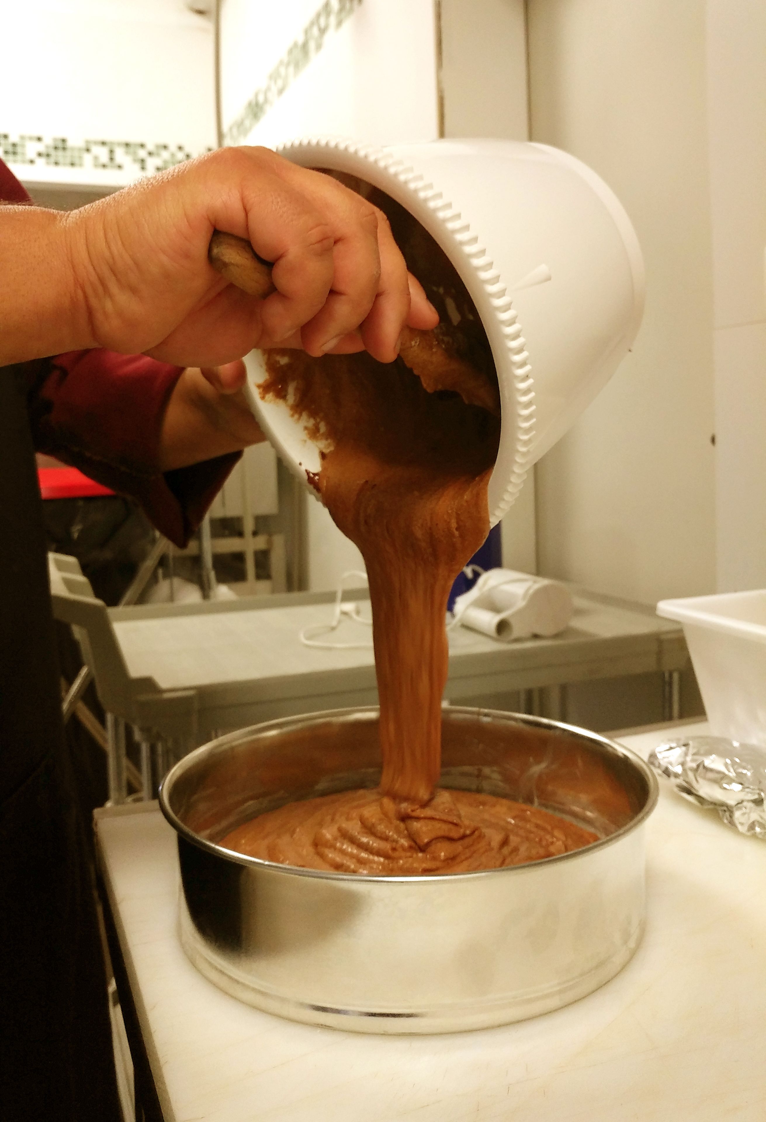 Une personne verse un liquide épais et brun d’un récipient blanc dans une poêle en métal, le tout posé sur un plan de travail blanc.