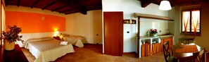 Una habitación con dos camas, una pequeña cocina con mesa y sillas, y un ventanal con cortinas.