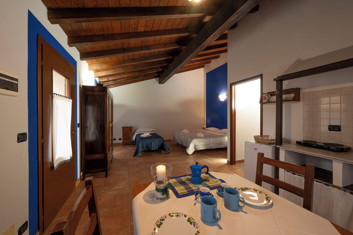 A room with a wooden ceiling, a bed, a table with a candle, and a small refrigerator.