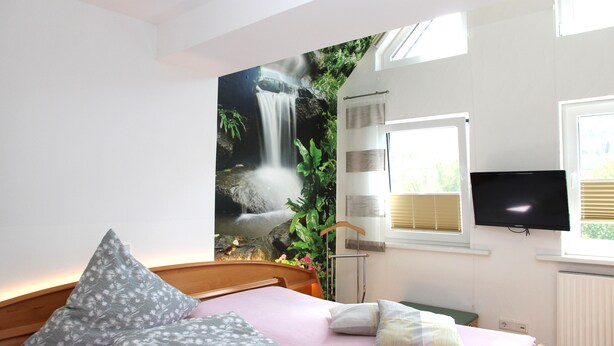A bedroom with a bed, pillows, a TV, and a wall mural of a waterfall. - Pension & Ferienwohnung Schweinsberg (Medebach)