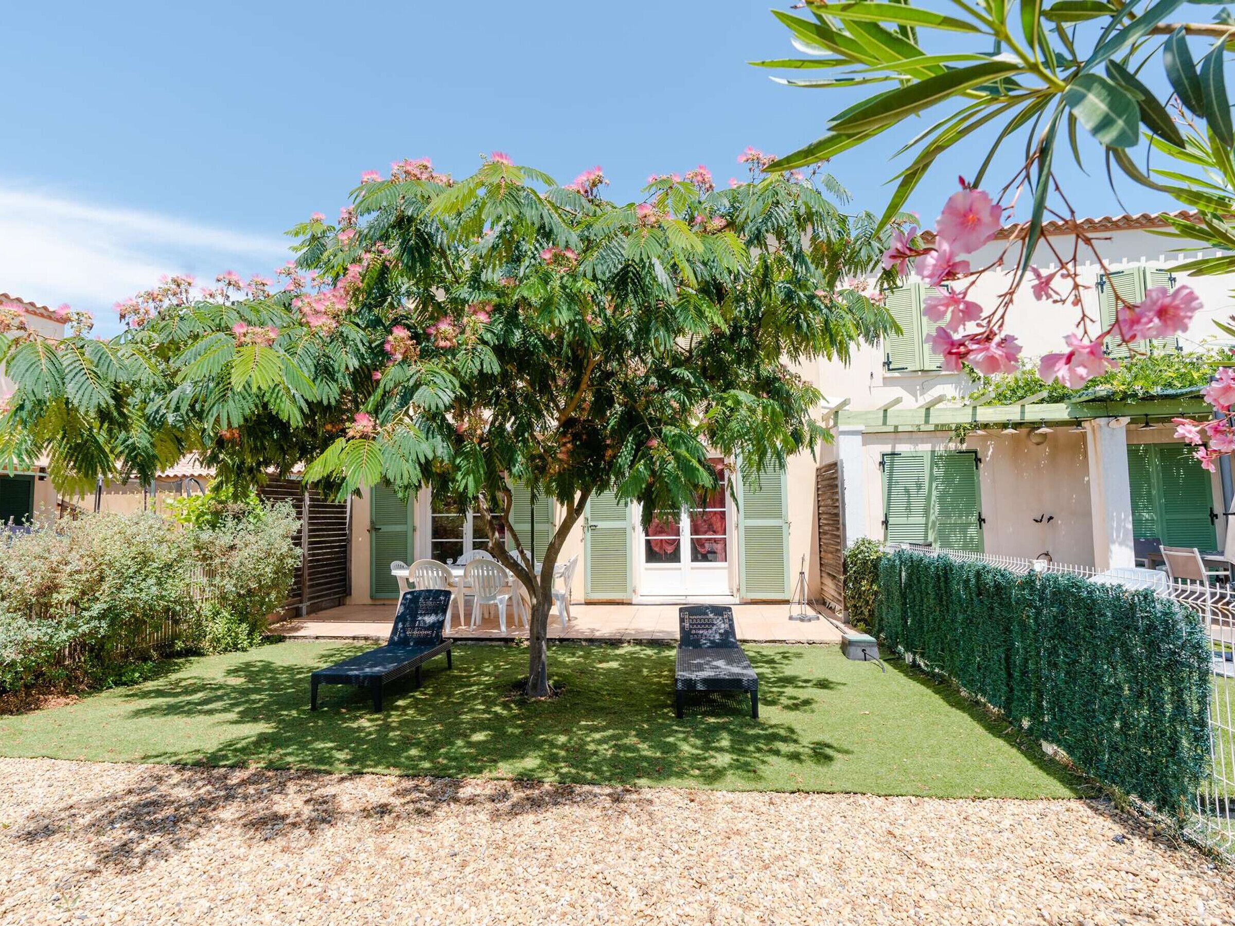 Un jardin avec deux chaises longues, un petit arbre aux fleurs roses et un bâtiment blanc aux volets verts.
