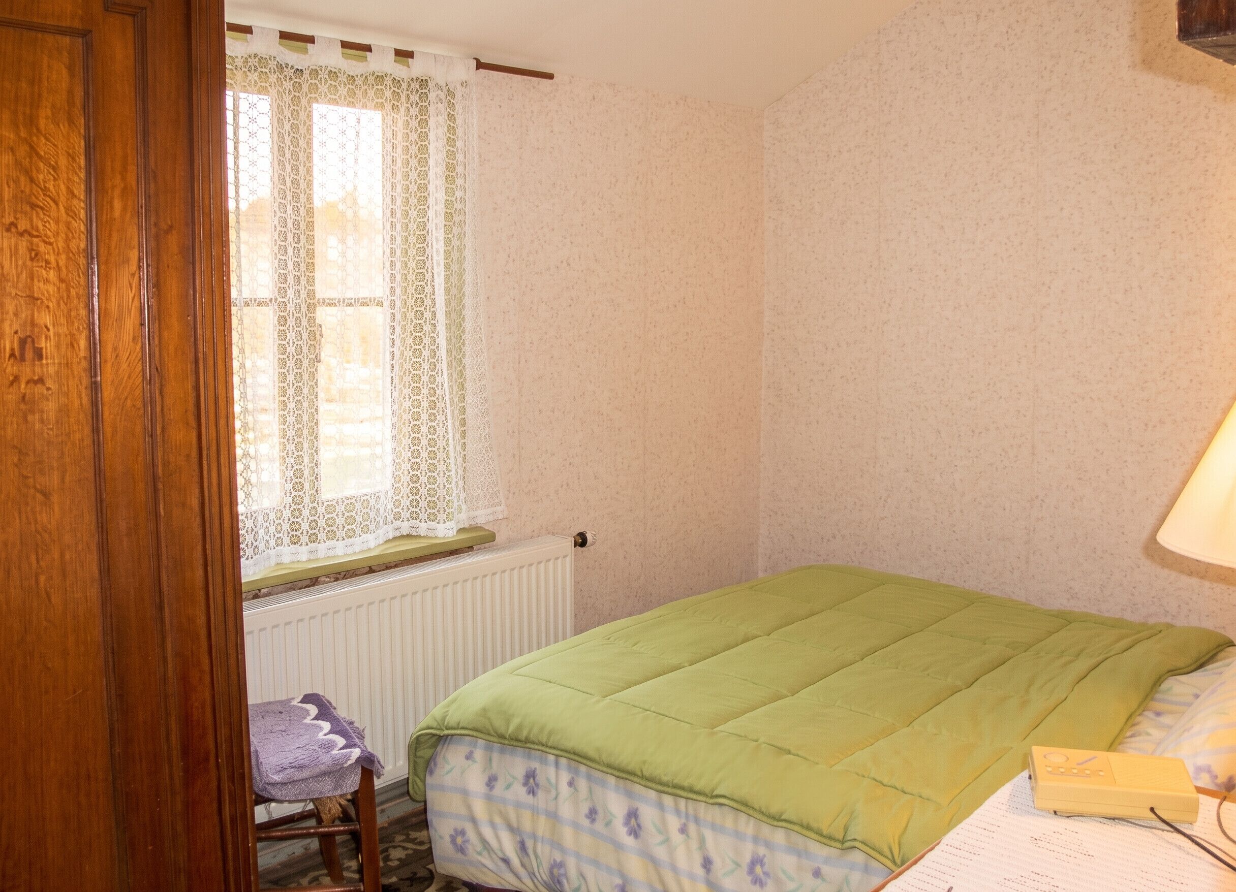 A bedroom with a bed, a window with lace curtains, a radiator, a chair with a purple towel, and a lamp on the bedside table.