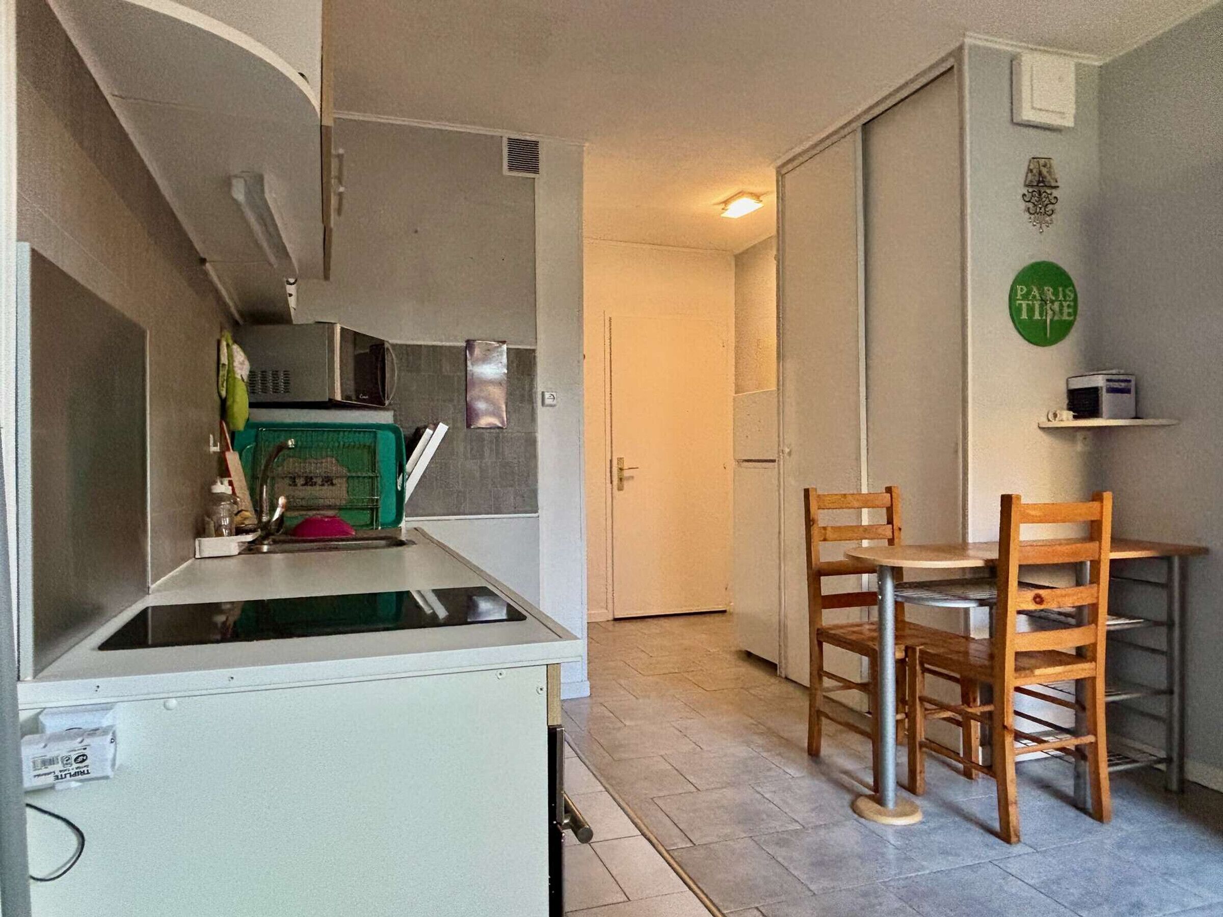 A compact kitchen with a microwave, sink, and green storage unit. There is a wooden chair and a small table in the adjacent dining area.