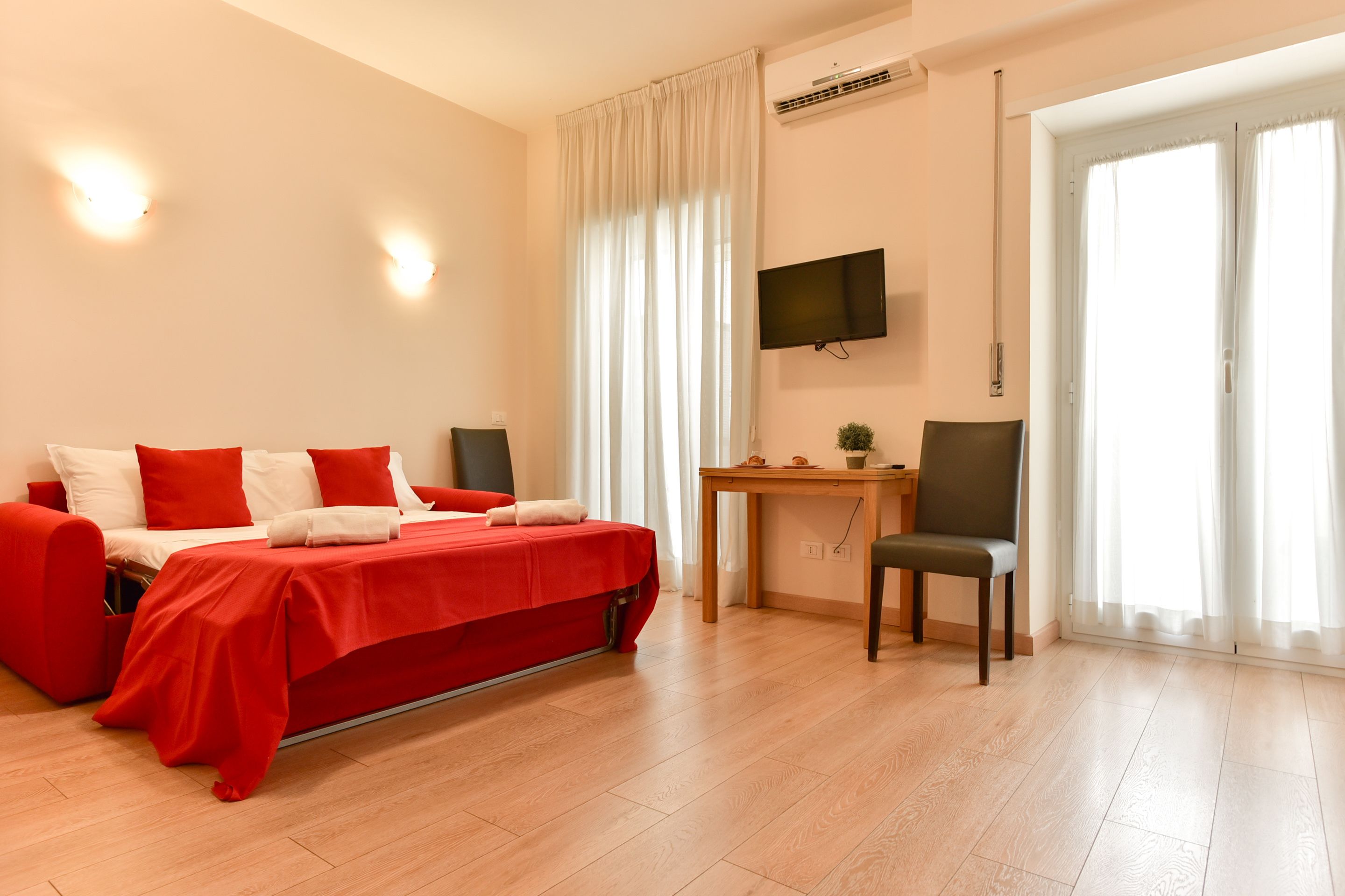 A modern hotel room with a red bed, a desk, a chair, and a TV on the wall.