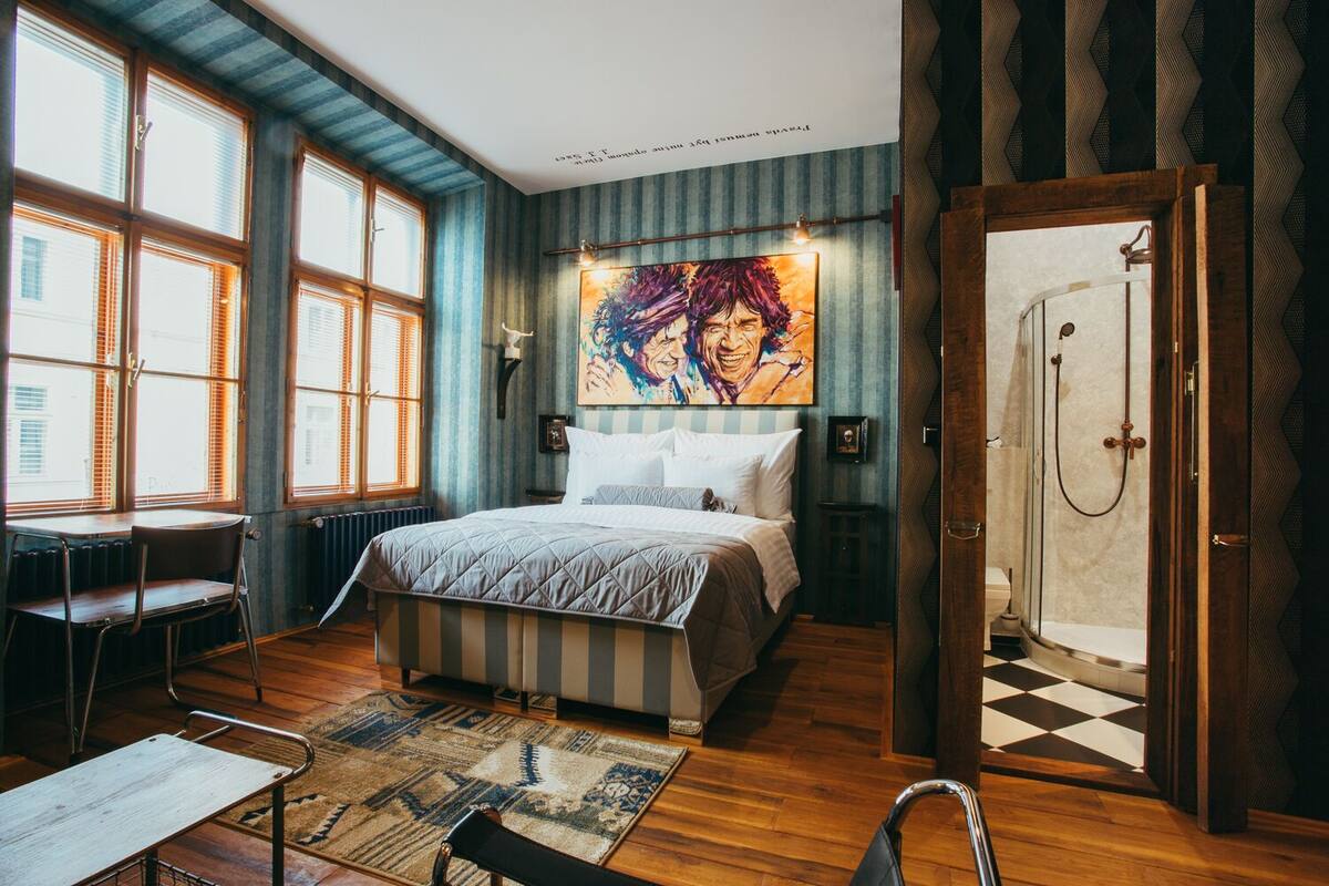 A bedroom with a large bed, a desk, and a chair, a shower, and a window with blinds.