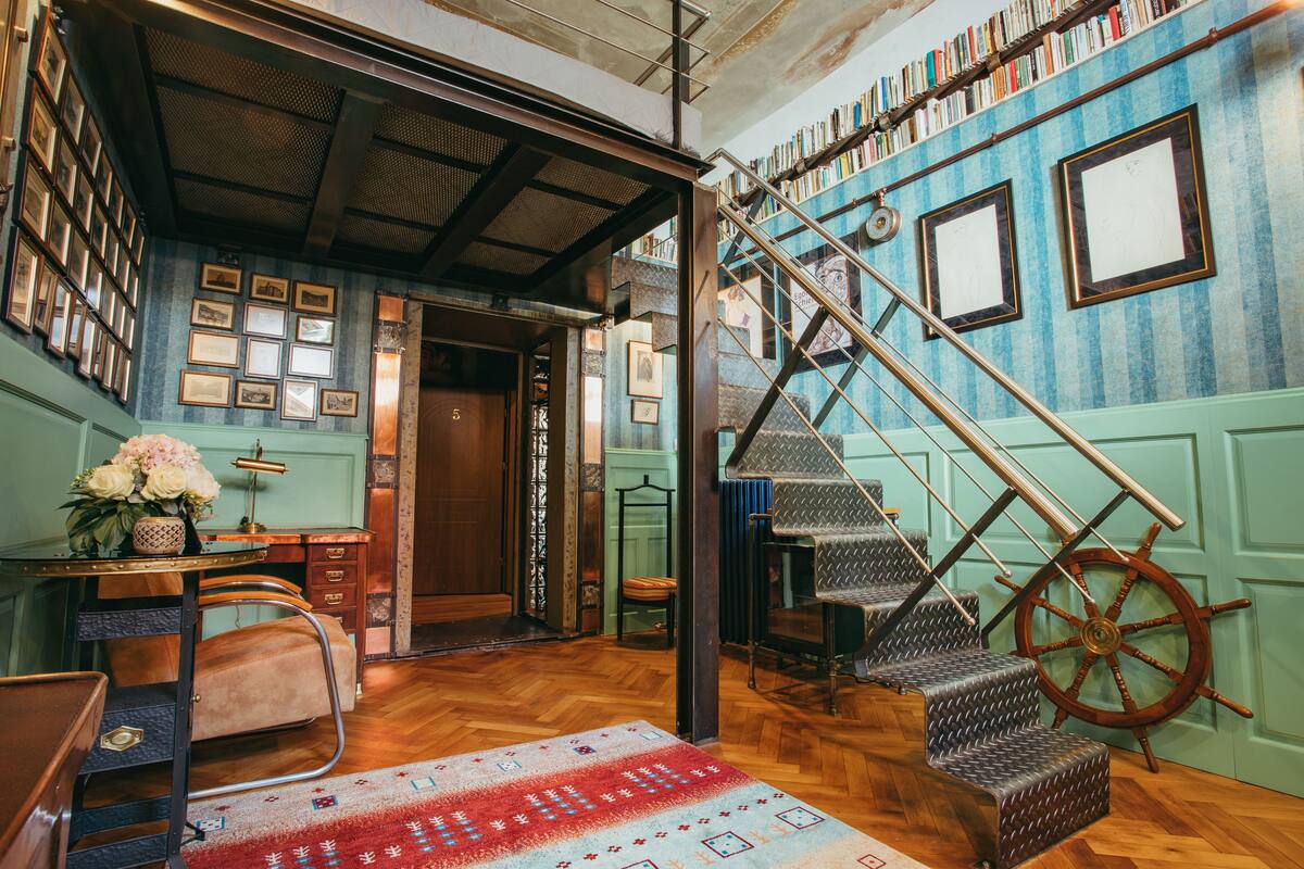 A room with a wooden desk, a chair, a staircase with a ship's wheel, and a wall adorned with framed pictures and a bookshelf.