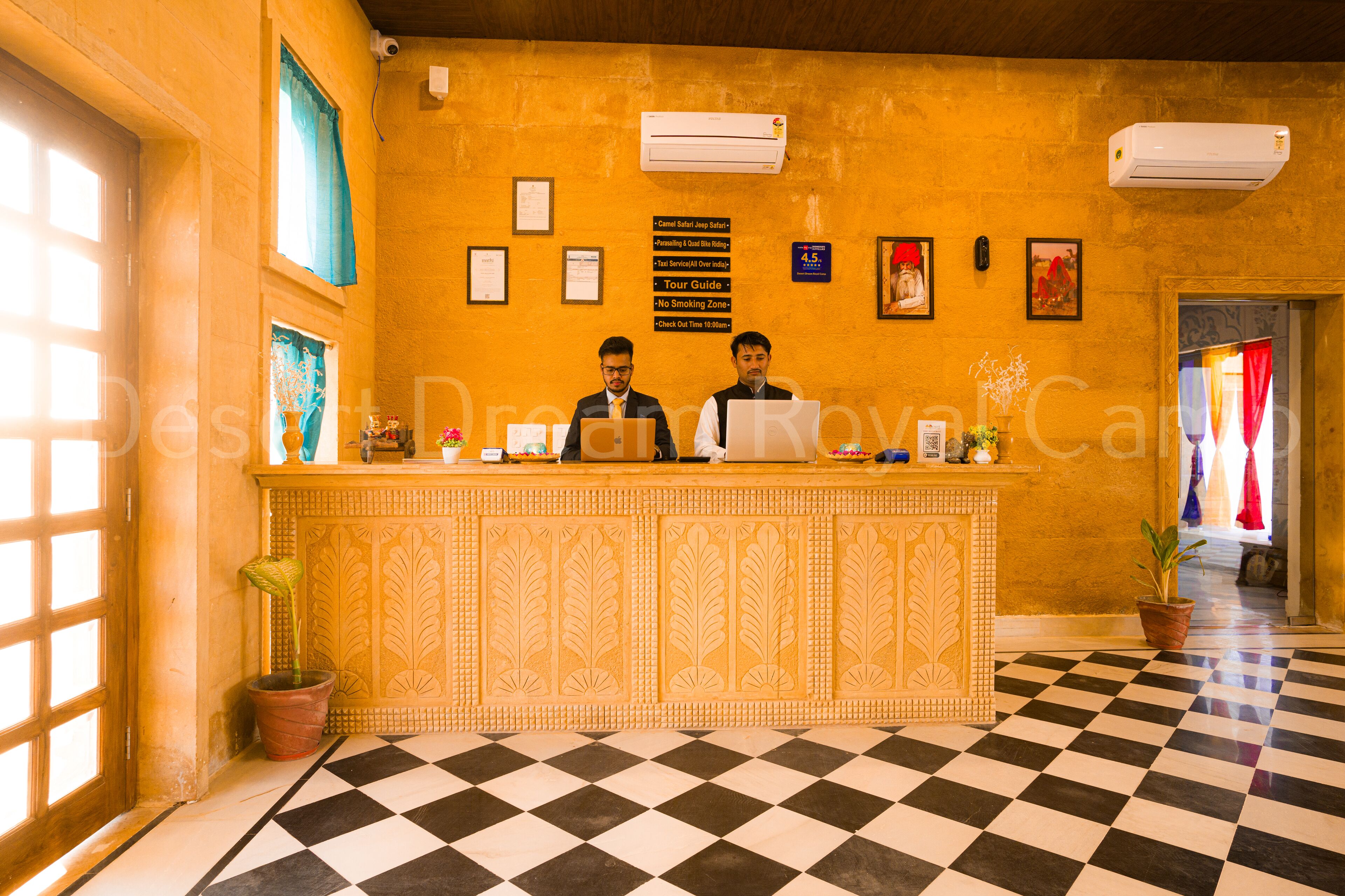A reception desk with two staff members, a check-in counter, and a check-out counter. There are framed certificates on the wall, a potted plant, and a black and white checkered floor.