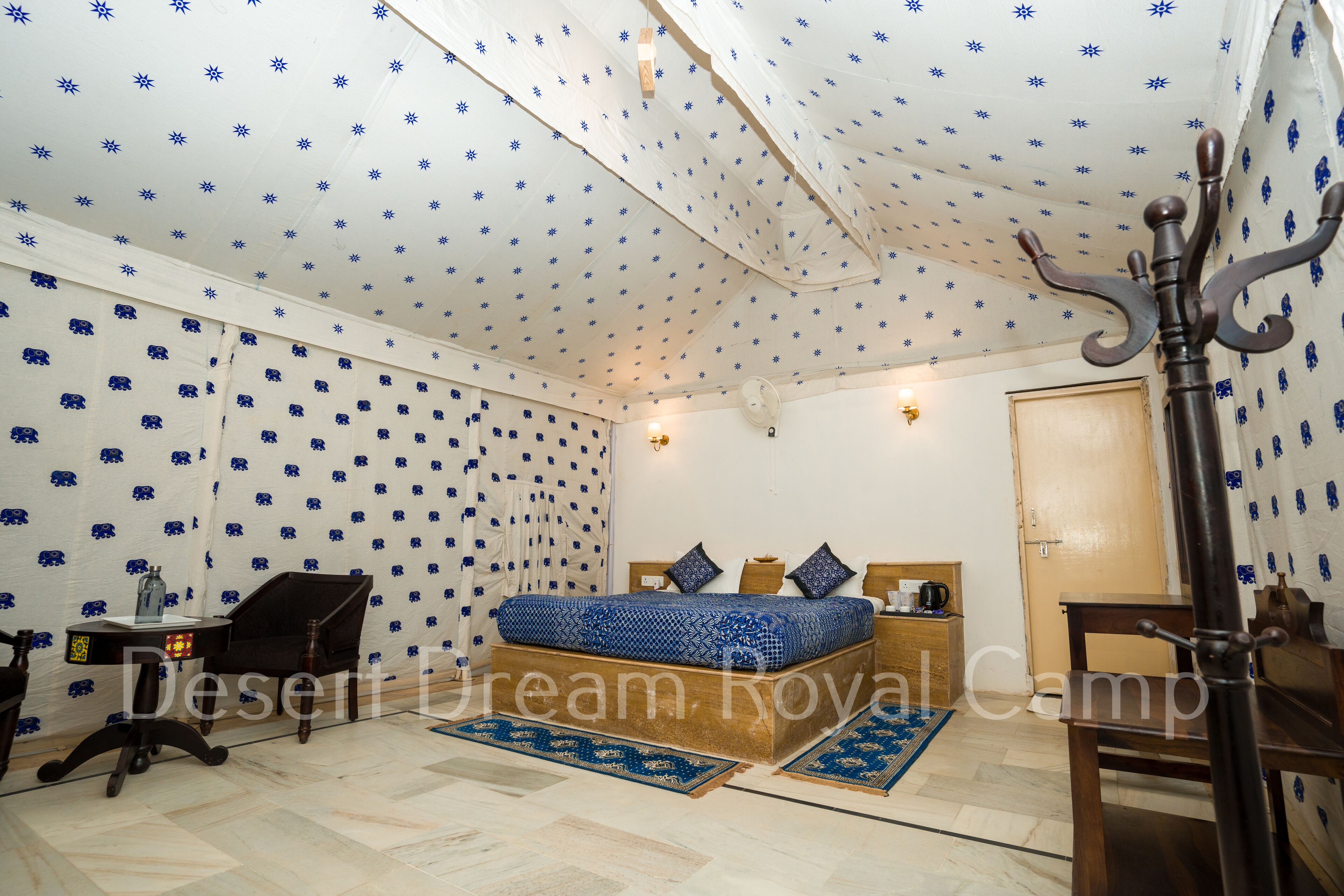 A room with a patterned ceiling, a bed with blue bedding, a wooden nightstand, a chair, and a desk with a lamp.