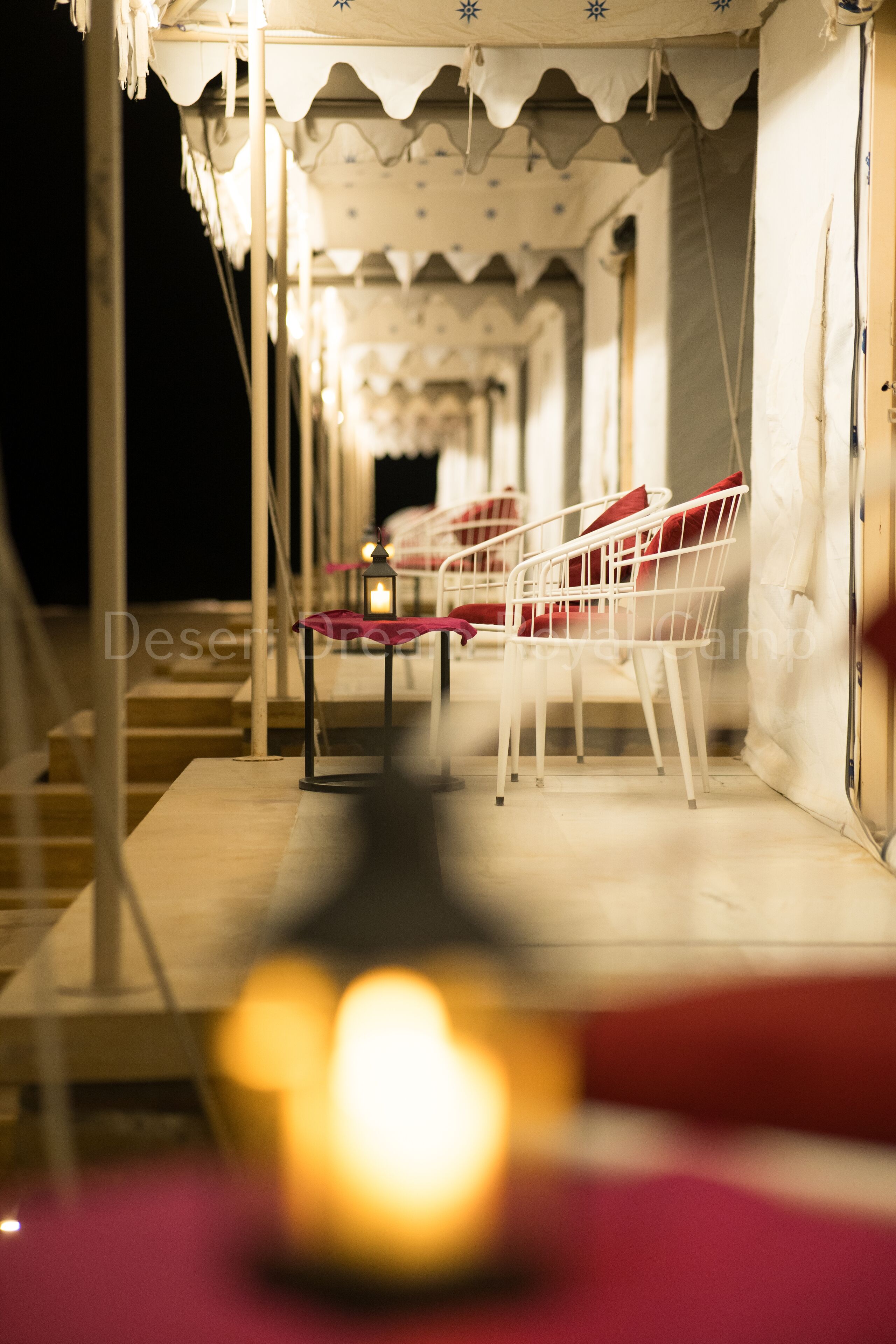 A corridor with white metal chairs and a table, a lit candle, and a tent-like structure with a patterned ceiling.