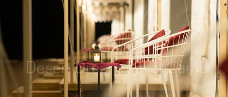 A corridor with white metal chairs and a table, a lit candle, and a tent-like structure with a patterned ceiling.