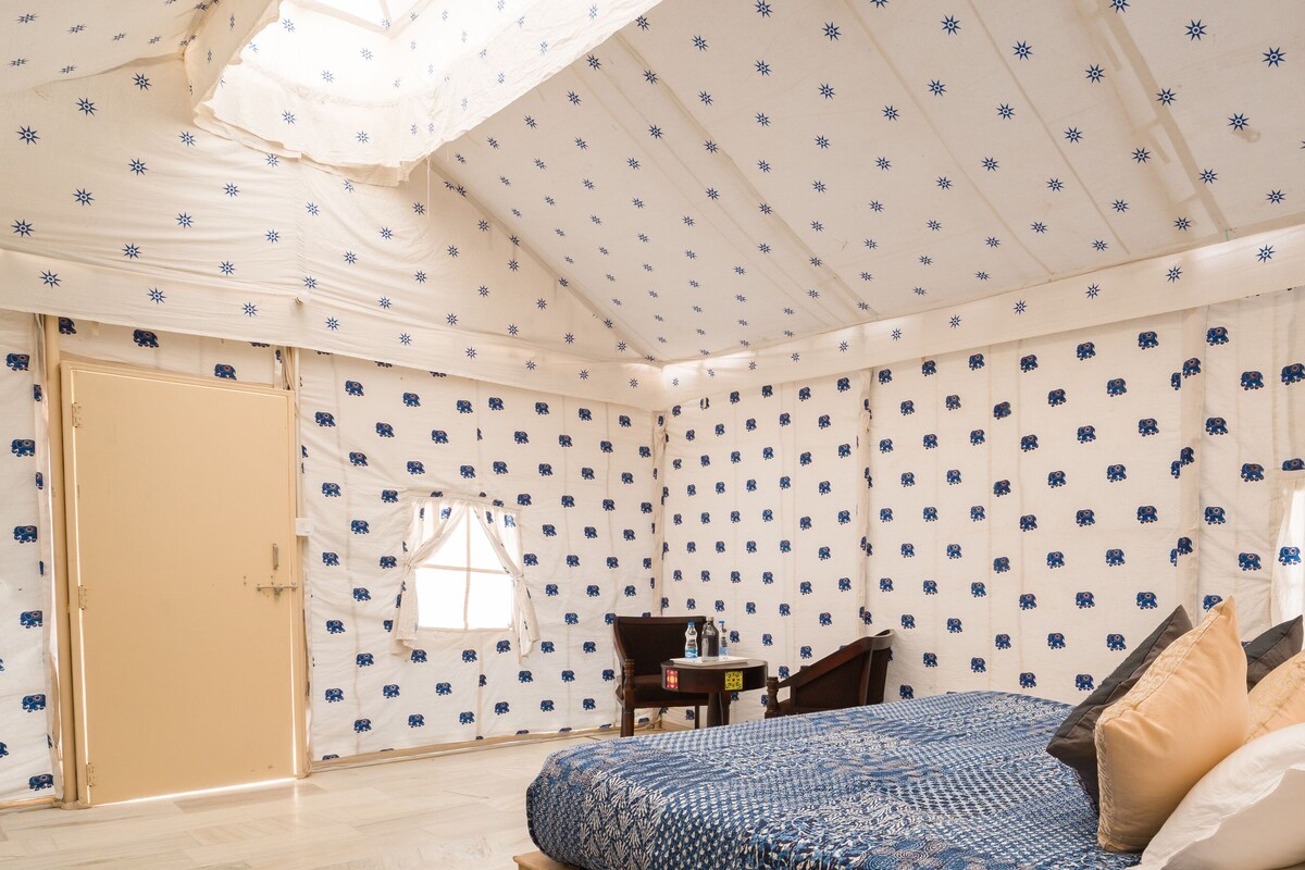 Una habitación con techo estampado, una cama con colcha azul y blanca, una mesita con una botella y una silla.