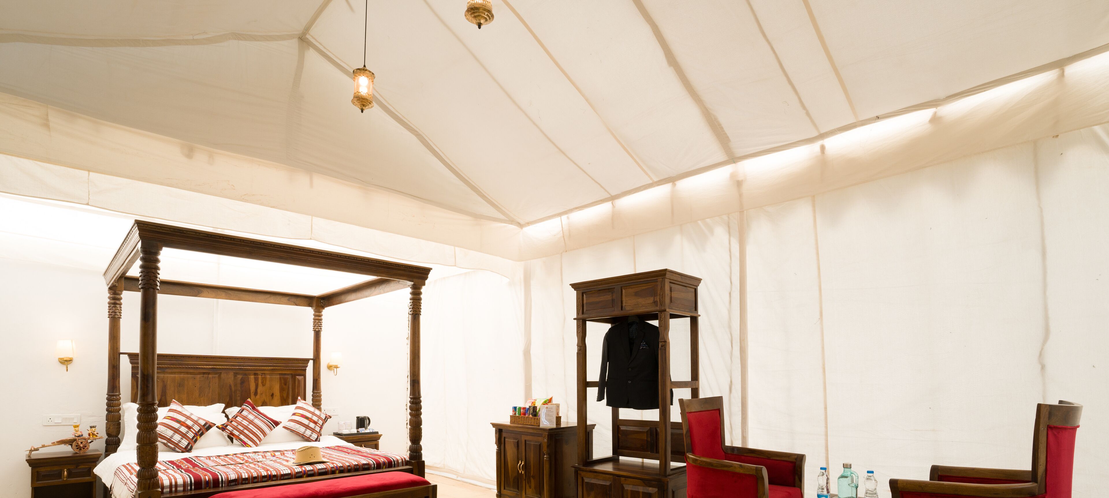 A four-poster bed with a red bedspread, a wooden nightstand with a lamp, and a wardrobe with a coat hanging inside.