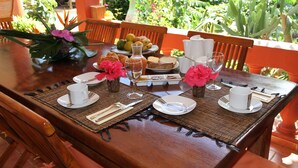 A dining table set with plates, cups, and cutlery, surrounded by chairs, with a view of lush greenery in the background. - Villa Maora (Koungou)