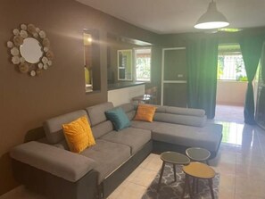 A living room with a sectional sofa, two small side tables, a mirror, and a view of the outdoors. - Villa Maora (Koungou)