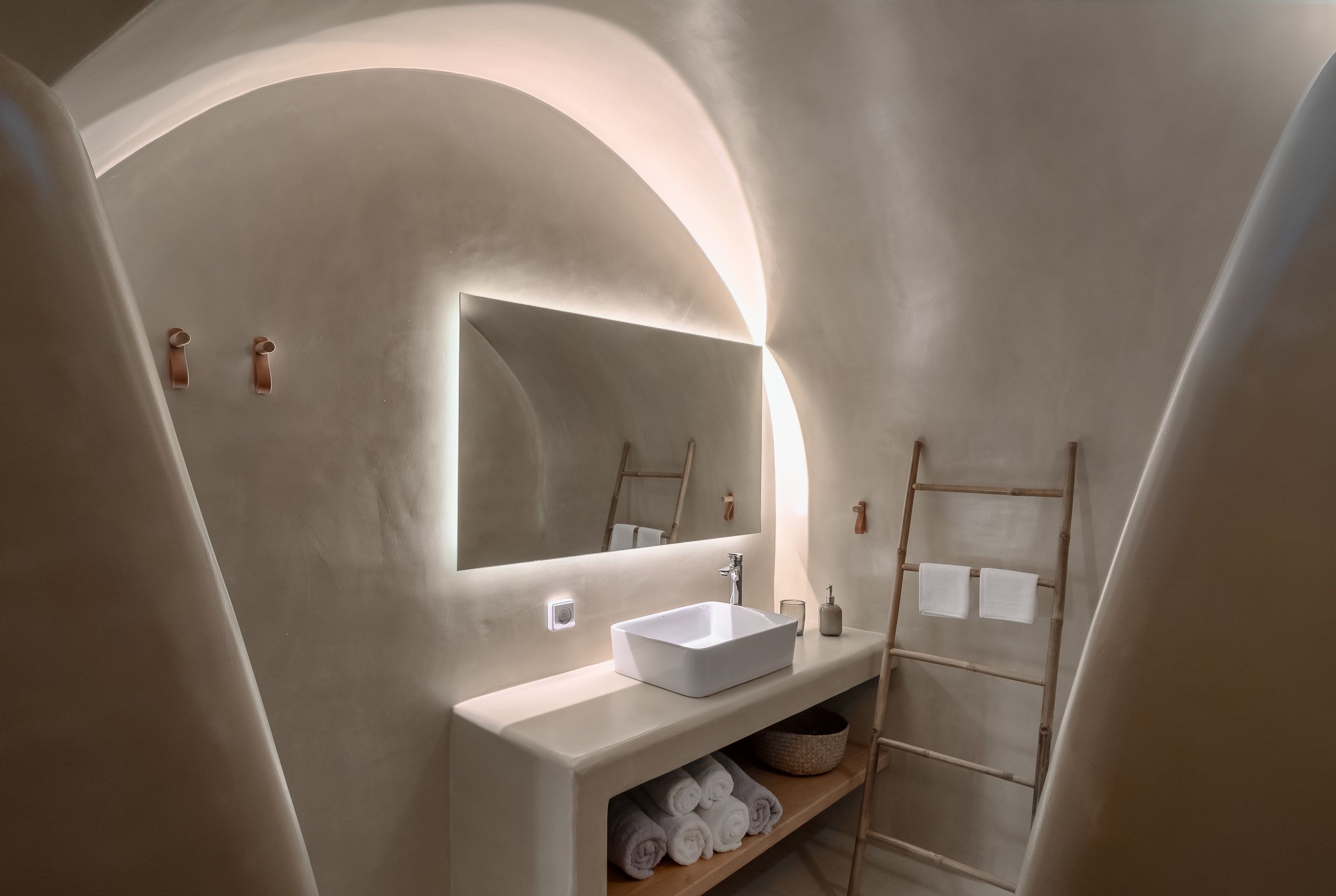 A modern bathroom with a white sink, a large mirror, and a wooden shelf with towels.