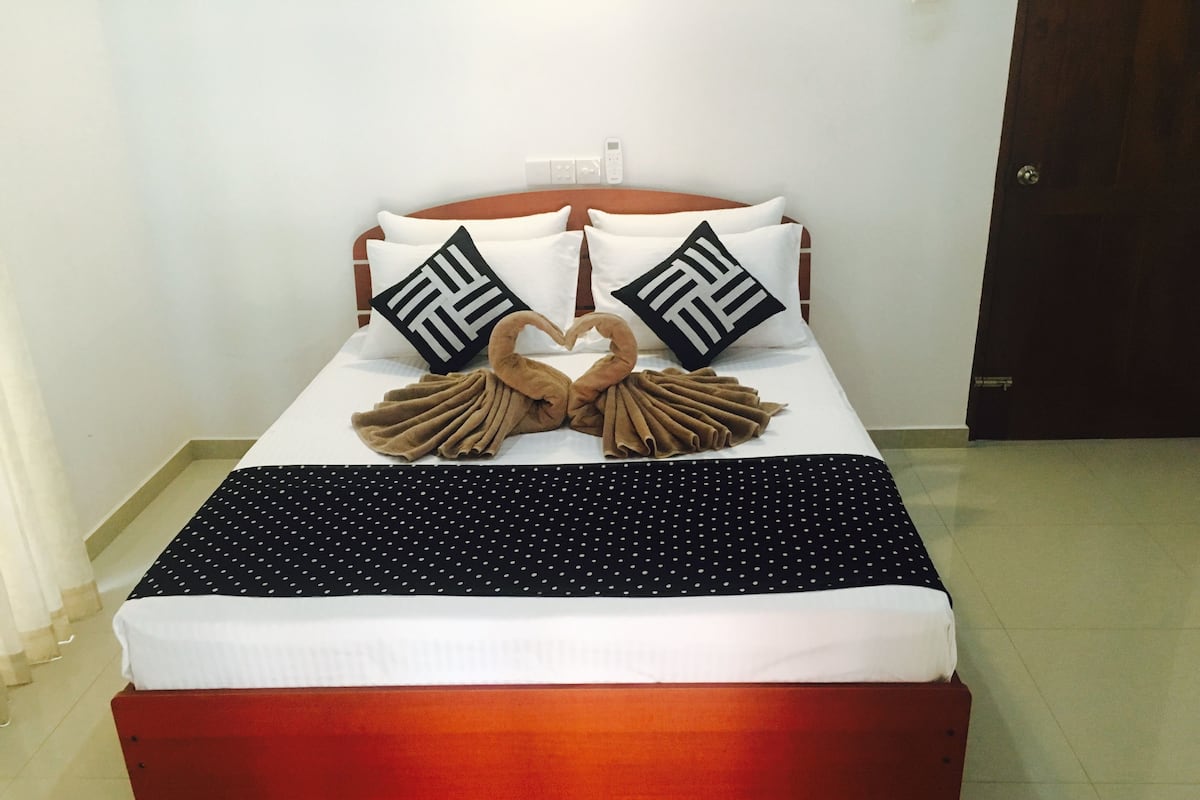 A neatly made bed with a heart-shaped towel arrangement, two pillows with a geometric pattern, and a polka-dotted blanket.