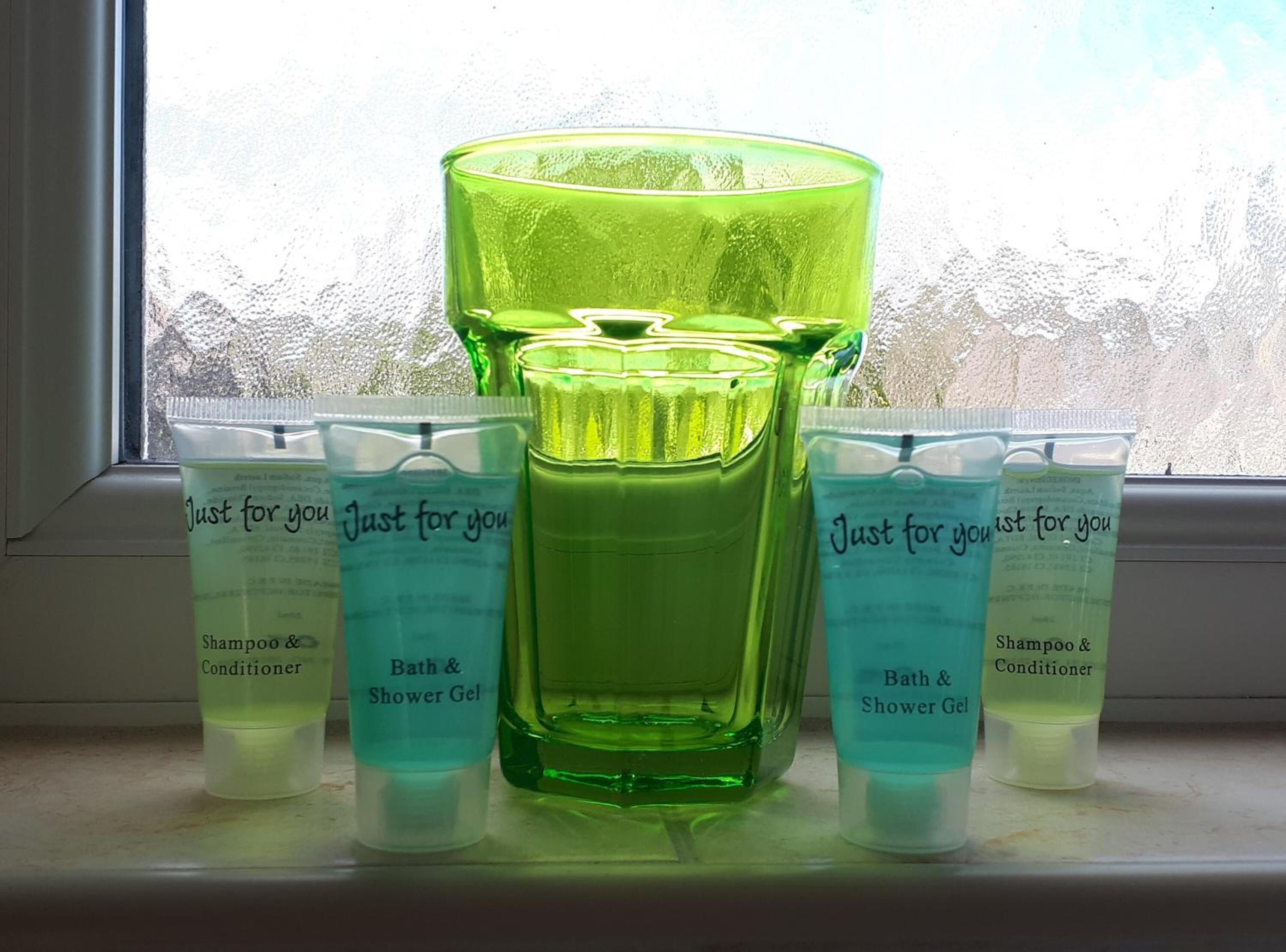 Estante de baño con botellas y tubos de vidrio verde etiquetados 'Just for you' que contienen champú, acondicionador y gel de ducha.