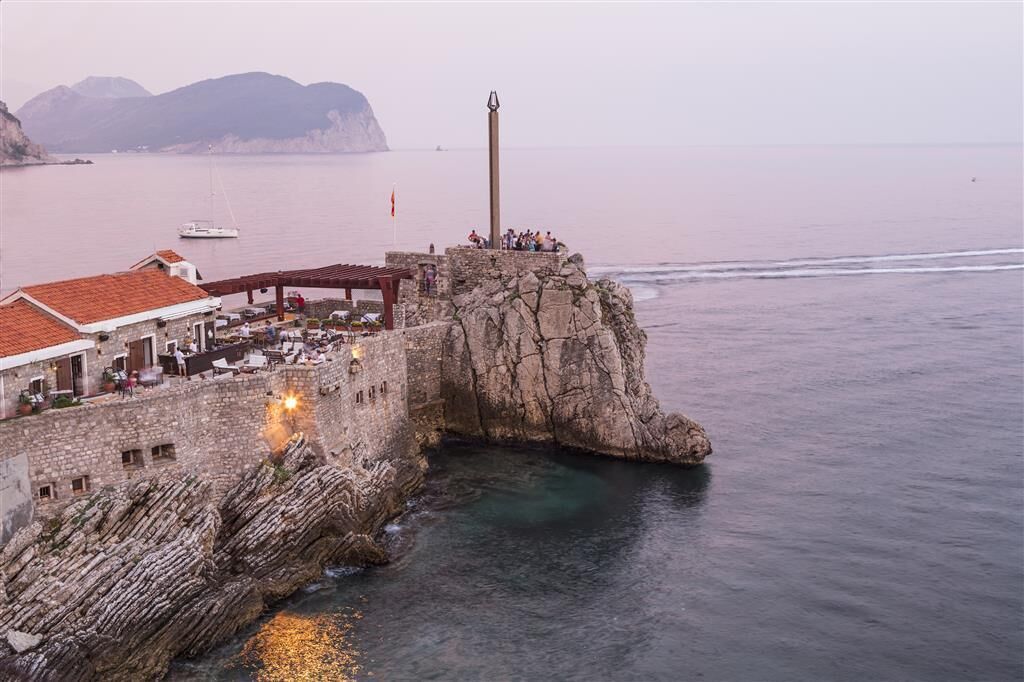 Una edificación costera de piedra con un faro, un muelle con asientos y un edificio de techo rojo.