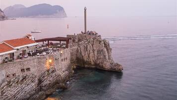 Una edificación costera de piedra con un faro, un muelle con asientos y un edificio de techo rojo.