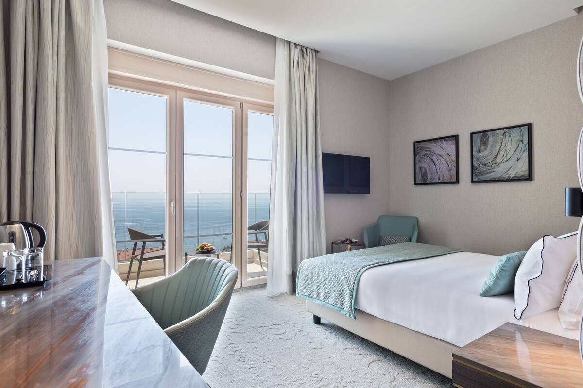 Habitación de hotel con cama, escritorio, silla, televisión y balcón con vistas al océano.