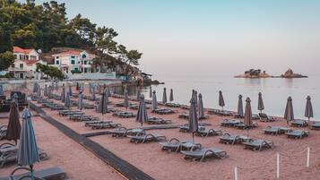 Ein Strand mit Reihen von Liegestühlen und Sonnenschirmen, einem hölzernen Steg und Gebäuden im Hintergrund.