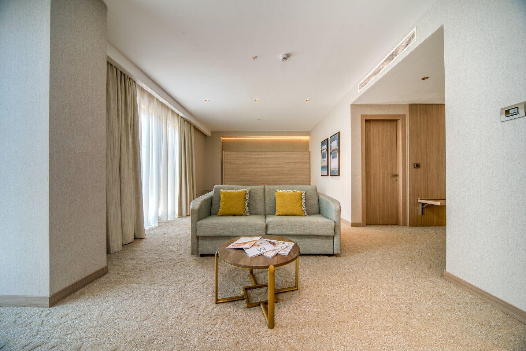 Quarto de hotel moderno com sofá, uma pequena mesa, porta de madeira e vista para o exterior.