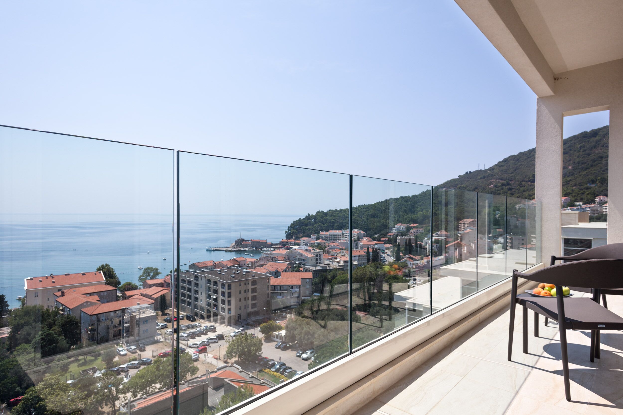 A balcony with a clear view of a coastal town, a dining area with chairs and a table, and a glass railing.