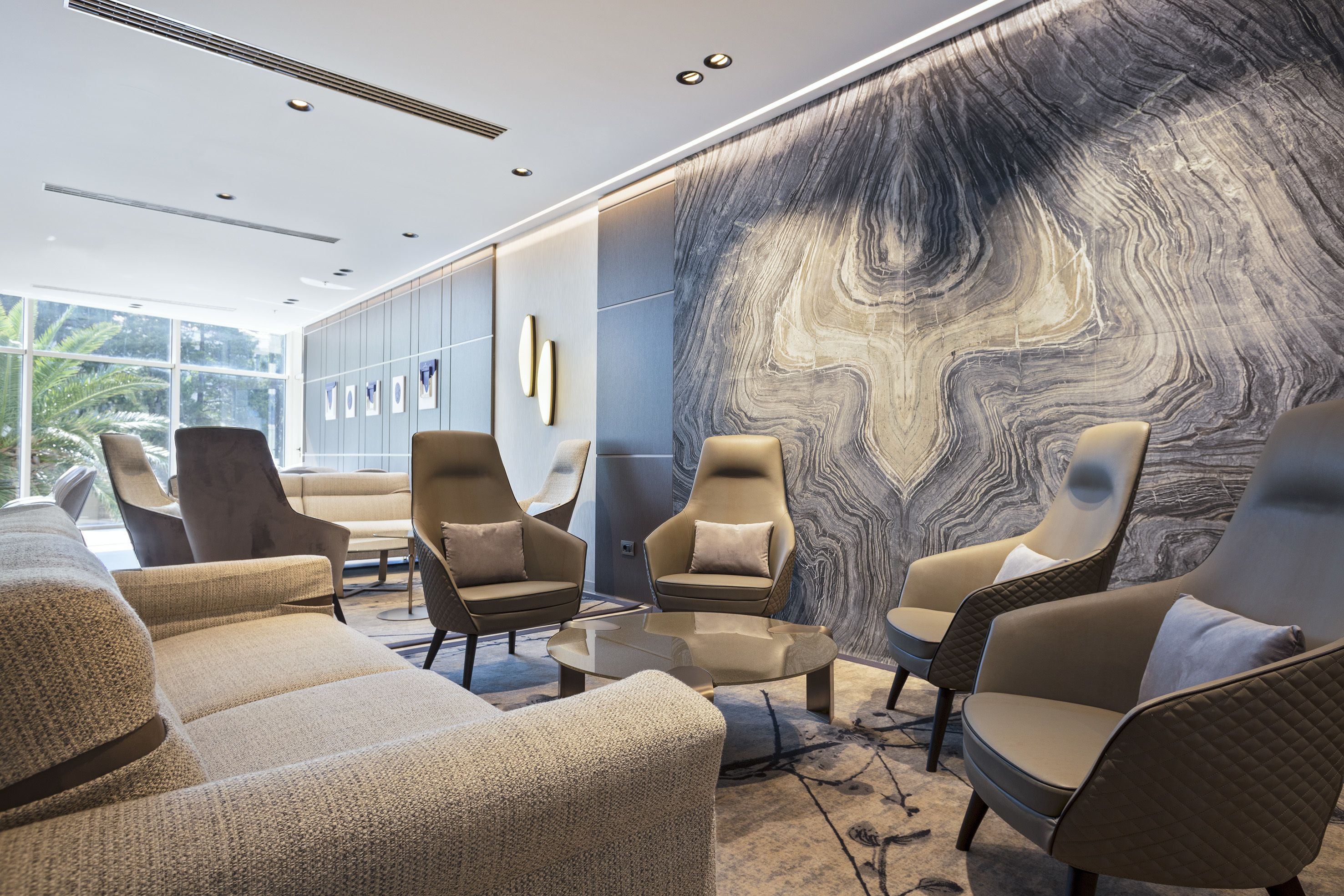 A modern lounge area with leather sofas and armchairs, a marble wall, and a glass coffee table.