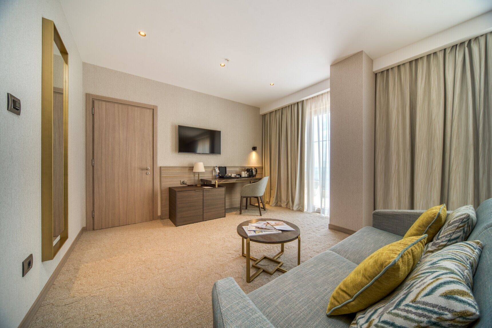 Una habitación de hotel con sofá, escritorio, televisor y un ventanal con cortinas.