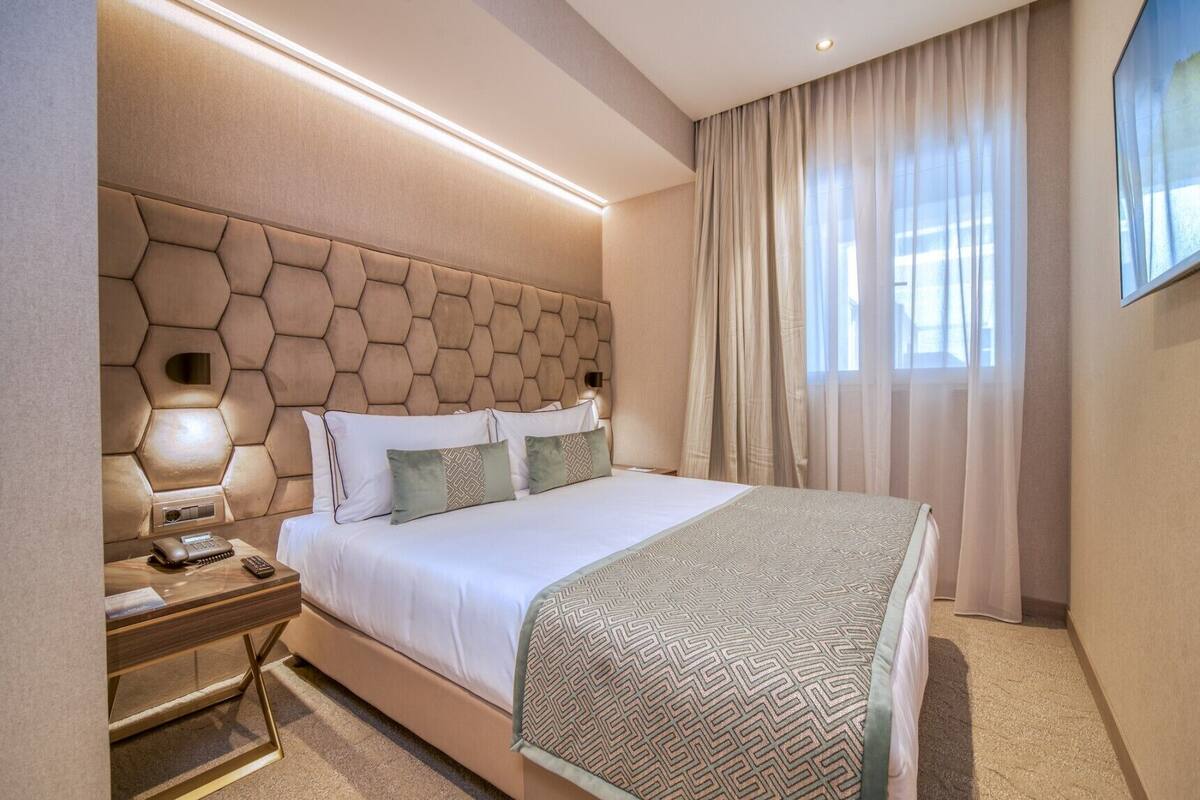 Habitación de hotel con cabecera hexagonal, cama con sábanas blancas, mesita de noche con lámpara y un ventanal con cortinas.