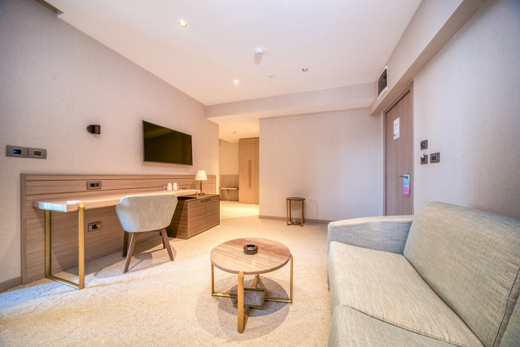 A modern hotel room with a flat-screen TV, a desk with a chair, a sofa, and a small round coffee table.