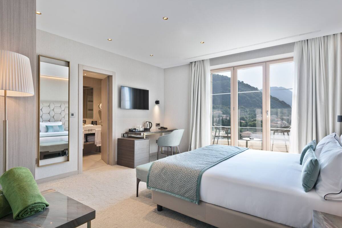 Habitación de hotel con cama, escritorio, silla, televisor y balcón con vista a la montaña.