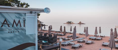 Une zone située au bord de la plage, avec une rangée de chaises longues et de parasols, un petit quai avec un bateau, et un panneau indiquant un hôtel, un centre de bien-être et un espace de conférence.
