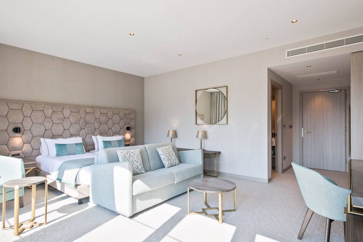Una habitación de hotel moderna con una cama grande, un sofá, una mesa redonda de centro y una silla. Hay un espejo en la pared y una puerta que da a otra habitación.